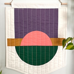 May include: Geometric wall hanging with a wooden dowel. The quilted fabric features a purple rectangle, a coral half-circle, a mustard-colored horizontal stripe, and a green half-circle. The background is off-white.