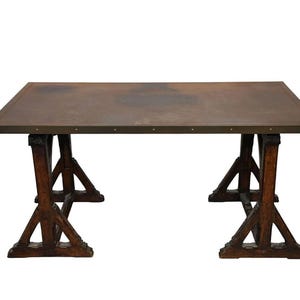 May include: A rectangular table featuring a flat, weathered copper-toned metal top with a dark patina and small metallic studs along the edges, supported by two dark brown wooden trestle-style bases with angled structural supports.