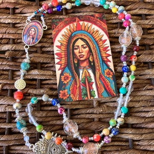 Puede incluir: Un rosario colorido con un colgante plateado con un diseño de ojo y una tarjeta con un retrato de una mujer con un halo. Las cuentas del rosario son multicolores, incluyendo cuentas claras, rojas, azules y verdes.