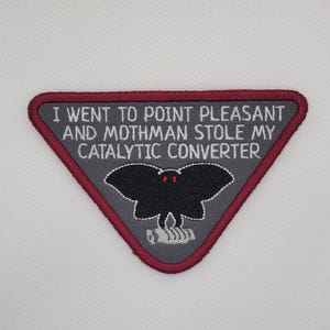 May include: A triangular patch with a grey background and a dark red border. The patch features the text "I WENT TO POINT PLEASANT AND MOTHMAN STOLE MY CATALYTIC CONVERTER." A black Mothman graphic is in the centre.