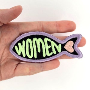 Könnte beinhalten: Ein gestickter Bügel-Patch in Form eines Fisches, in Lila und Schwarz mit dem Wort "FRAUEN" in grünen Buchstaben und einem rosa Herz auf der Schwanzflosse.