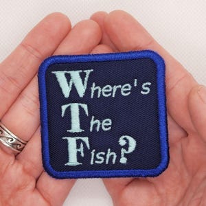 May include: A blue square patch with white and light blue lettering that reads "Where's The Fish?" The patch has a blue border.
