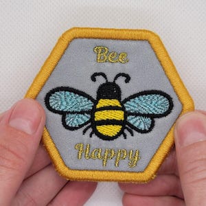 May include: A grey hexagonal embroidered patch with a yellow and black bee and the text "Bee Happy" in gold thread.