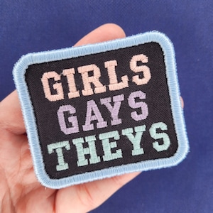 Könnte beinhalten: Ein schwarzer Stoff-Patch mit blauem Rand. Der Patch ist mit den Worten "GIRLS GAYS THEYS" in Pastellrosa, Lila und Grün bestickt.