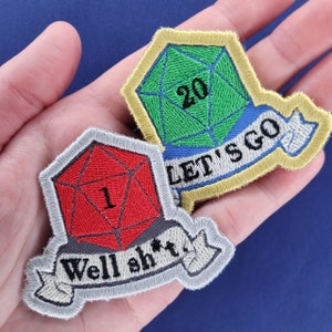 May include: Two embroidered patches featuring dice with a banner. The first patch is green and says "Let's Go" with a 20-sided die. The second patch is red and says "Well sh*t" with a 1-sided die.