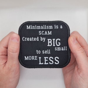 May include: Black patch with white text that reads "Minimalism is a SCAM Created by BIG to sell small MORE LESS".