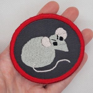 Könnte beinhalten: Ein runder, grauer gestickter Aufnäher mit rotem Rand. Der Aufnäher zeigt eine weiße Maus mit schwarzen Augen und einer rosa Nase.