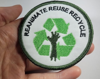 Puede incluir: Un parche circular blanco con un símbolo de reciclaje verde y una mano negra levantada. El texto "REANIMATE REUSE RECYCLE" está escrito en negro alrededor del símbolo.