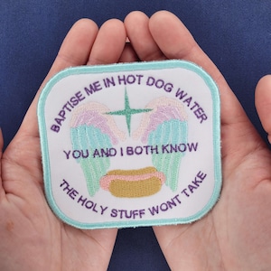 May include: Embroidered patch with the text "BAPTISE ME IN HOT DOG WATER YOU AND I BOTH KNOW THE HOLY STUFF WONT TAKE". The patch features angel wings, a cross, and a hot dog design, with a light blue border.