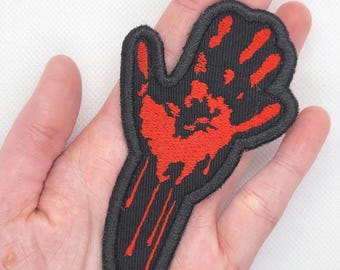 Puede incluir: Un parche bordado en negro y rojo de una huella de mano ensangrentada. La mano está extendida con cinco dedos y gotea sangre.