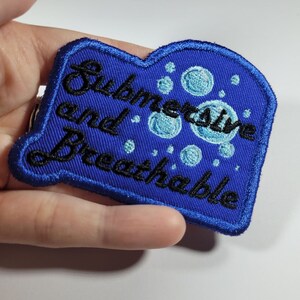 May include: Blue embroidered patch with the text "Submersive and Breathable" in black cursive font. The patch has a blue background with white and light blue bubbles.