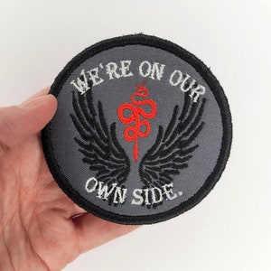 May include: A gray round embroidered patch with black wings and a red snake with a staff. The text on the patch reads "We're on our own side."