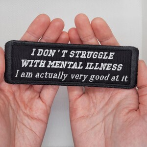 May include: A black fabric patch with white text that reads "I don't struggle with mental illness I am actually very good at it."