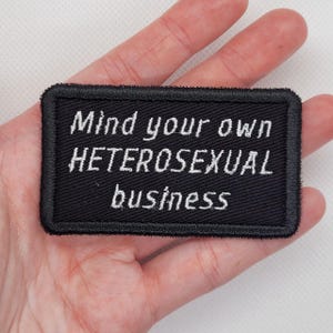 May include: A black rectangular patch with white embroidered text that reads "Mind your own HETEROSEXUAL business". The patch has a black border and is held in a hand.