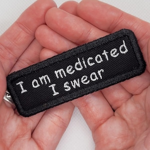 Könnte beinhalten: Schwarzer rechteckiger Aufnäher mit weiß gesticktem Text „I am medicated I swear“. Der Aufnäher hat einen schwarzen Rand und wird in offenen Händen gehalten.