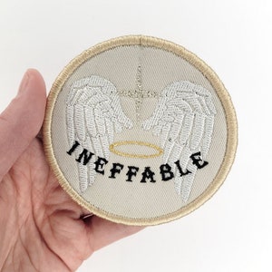 May include: Round embroidered patch with a beige background, featuring white angel wings, a gold halo, and a cross. The word "INEFFABLE" is embroidered in black. The patch has a gold border.