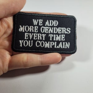 Könnte beinhalten: Ein schwarzer rechteckiger Aufnäher mit weiß gesticktem Text, der lautet: "WE ADD MORE GENDERS EVERY TIME YOU COMPLAIN." Der Aufnäher hat einen schwarzen Rand und wird in einer Hand gehalten.