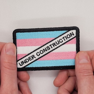 May include: A blue, pink and white transgender pride flag patch with black text that reads "UNDER CONSTRUCTION" across the flag.