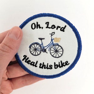 May include: A white round embroidered patch with a blue border. The patch features a black and white illustration of a bicycle with a basket and the text "Oh, Lord Heal this bike".