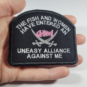 May include: Black embroidered patch with white text. The text reads "THE FISH AND WOMEN HAVE ENTERED AN UNEASY ALLIANCE AGAINST ME." The patch features a pink fish skeleton and crossed silver swords.