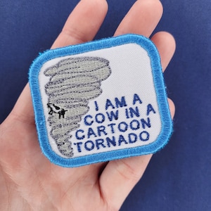 May include: A blue and white embroidered patch with a cartoon tornado and the text "I am a cow in a cartoon tornado".