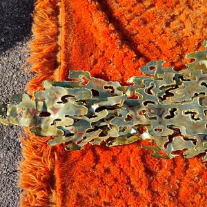 May include: A gold-toned metal wall sculpture with a textured, organic design. The sculpture has a flowing, abstract shape and appears to be made of multiple layers of metal.