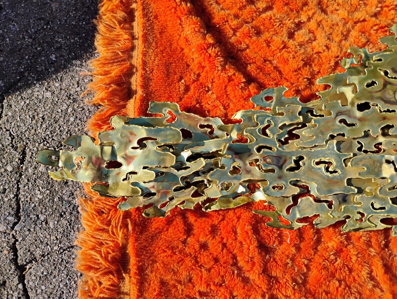 May include: A gold-toned metal wall sculpture with a textured, organic design. The sculpture has a flowing, abstract shape and appears to be made of multiple layers of metal.