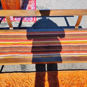 May include: A mid-century modern bench with a wooden frame and a striped fabric seat in shades of brown, orange, yellow, and red. The bench has a backrest and angled legs, showcasing a retro design.