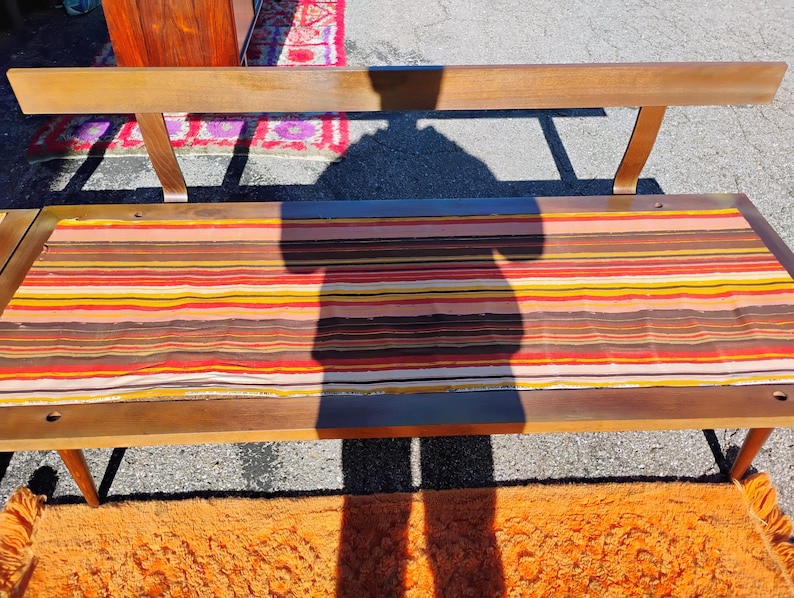 May include: A mid-century modern bench with a wooden frame and a striped fabric seat in shades of brown, orange, yellow, and red. The bench has a backrest and angled legs, showcasing a retro design.