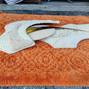 May include: An abstract sculpture with a cream-coloured, textured surface and a gold-coloured metal accent. The sculpture is set on a vibrant orange shag rug, creating a striking contrast in textures and colours.