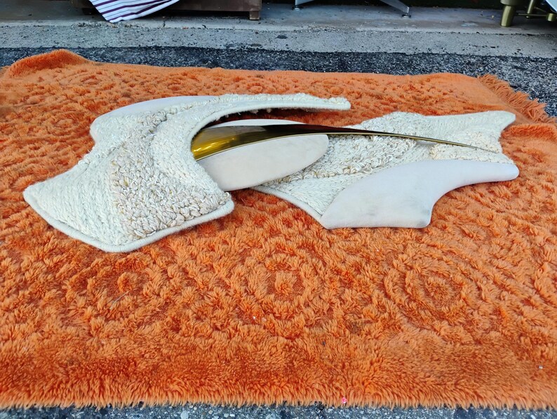 May include: An abstract sculpture with a cream-coloured, textured surface and a gold-coloured metal accent. The sculpture is set on a vibrant orange shag rug, creating a striking contrast in textures and colours.