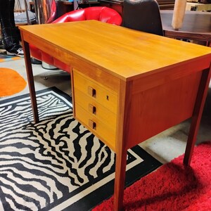 May include: A wooden desk with a light brown finish. The desk has a rectangular top and three drawers on the right side. The desk is supported by four slender legs. The desk is placed on a zebra-striped rug.