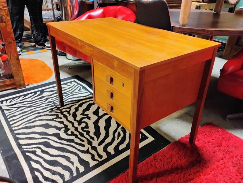 May include: A wooden desk with a light brown finish. The desk has a rectangular top and three drawers on the right side. The desk is supported by four slender legs. The desk is placed on a zebra-striped rug.