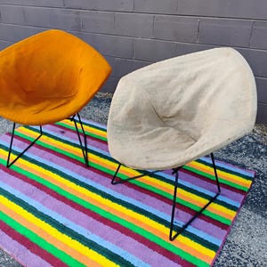 May include: Two diamond-shaped chairs with black metal wire frames. One chair features a vibrant orange fabric seat, while the other has a light beige fabric seat. Both chairs have a curved, shell-like design and are positioned on a striped rug.