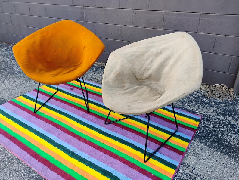 May include: Two diamond-shaped chairs with black metal wire frames. One chair features a vibrant orange fabric seat, while the other has a light beige fabric seat. Both chairs have a curved, shell-like design and are positioned on a striped rug.