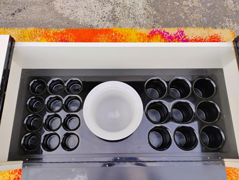 May include: A black bar cart with a white top and a black interior. The interior has a large white bowl and 12 small black cups.
