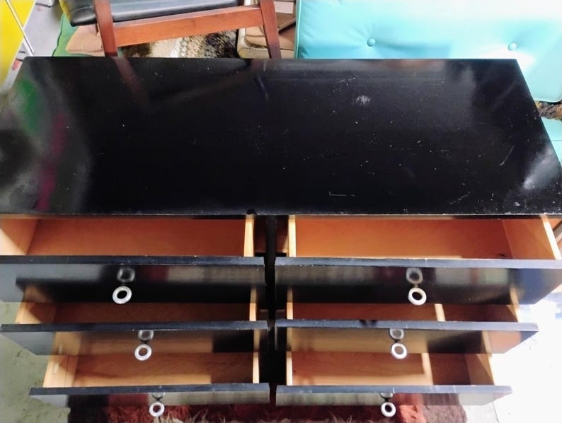 May include: A black dresser with six drawers, each with a white circular handle. The drawers are open, revealing a light brown interior. The top of the dresser is glossy black.