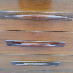 May include: A wooden chest of drawers featuring three horizontal drawers with a smooth, polished finish. Each drawer is equipped with a long, tapered, dark brown wooden handle that spans the center, providing a sleek and uniform appearance.