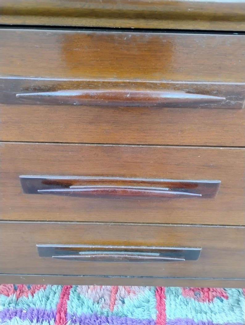 May include: A wooden chest of drawers featuring three horizontal drawers with a smooth, polished finish. Each drawer is equipped with a long, tapered, dark brown wooden handle that spans the center, providing a sleek and uniform appearance.