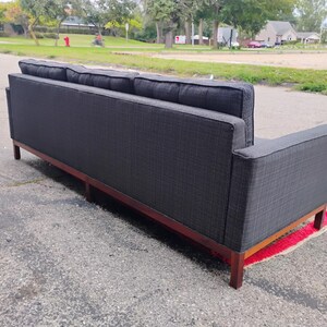 K&ouml;nnte beinhalten: Ein anthrazitfarbenes Sofa mit strukturiertem Stoffbezug, gerader R&uuml;ckenlehne, quadratischen Armlehnen und einem dunkelbraunen Holzsockel mit vier Beinen. Das Sofa verf&uuml;gt &uuml;ber drei R&uuml;ckenkissen und ein langes, durchgehendes Sitzpolster.
