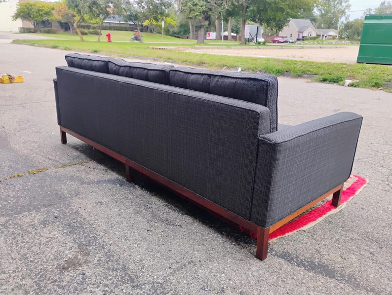 K&ouml;nnte beinhalten: Ein anthrazitfarbenes Sofa mit strukturiertem Stoffbezug, gerader R&uuml;ckenlehne, quadratischen Armlehnen und einem dunkelbraunen Holzsockel mit vier Beinen. Das Sofa verf&uuml;gt &uuml;ber drei R&uuml;ckenkissen und ein langes, durchgehendes Sitzpolster.