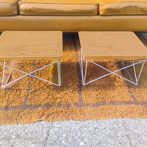 May include: A pair of square side tables featuring light brown wood grain laminate tops and white wire metal bases with a crisscross structural design. The tables have rounded corners and are positioned side by side on a textured surface.
