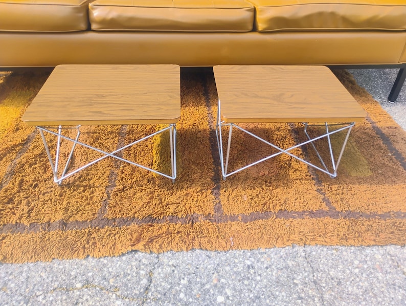 May include: A pair of square side tables featuring light brown wood grain laminate tops and white wire metal bases with a crisscross structural design. The tables have rounded corners and are positioned side by side on a textured surface.