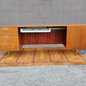 May include: A long wooden desk with a warm brown finish, featuring two stacked drawers on the left side with silver-toned handles, a central pull-out keyboard tray, and a single cabinet door on the right side, all supported by thin metal legs.