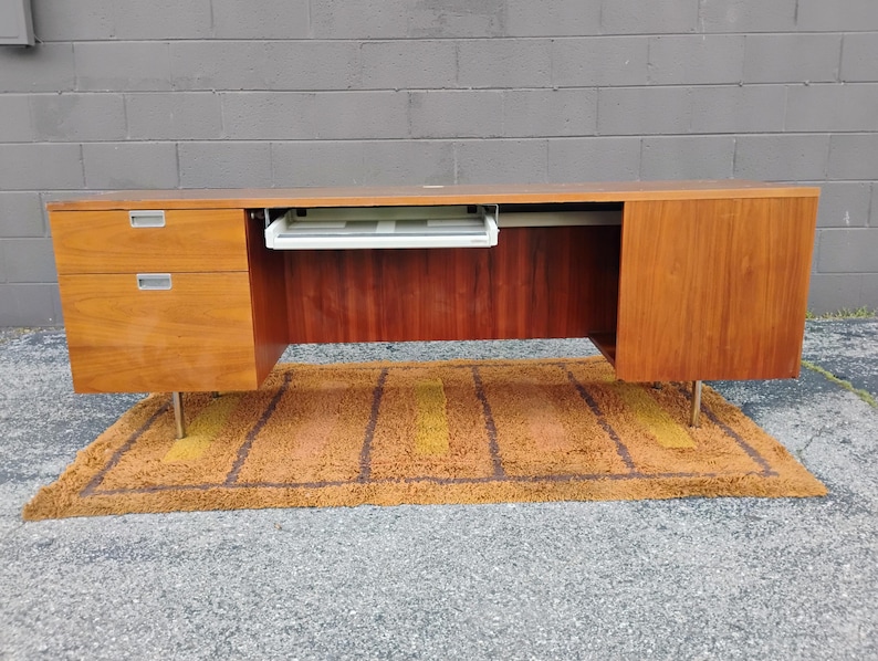 May include: A long wooden desk with a warm brown finish, featuring two stacked drawers on the left side with silver-toned handles, a central pull-out keyboard tray, and a single cabinet door on the right side, all supported by thin metal legs.