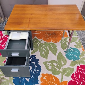 May include: A rectangular desk with a smooth, light brown wooden top and a grey metal base. The desk features two stacked drawers on the left side, both of which are pulled open. The top drawer contains a white plastic organiser tray with compartments.
