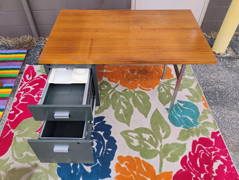 May include: A rectangular desk with a smooth, light brown wooden top and a grey metal base. The desk features two stacked drawers on the left side, both of which are pulled open. The top drawer contains a white plastic organiser tray with compartments.