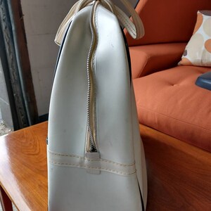 Pode incluir: Bolsa de viagem de vinil branco com detalhes pretos e fecho de correr. A bolsa tem uma al&ccedil;a superior e est&aacute; sobre uma superf&iacute;cie de madeira.