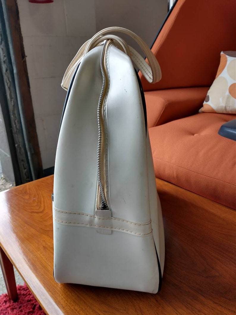 Pode incluir: Bolsa de viagem de vinil branco com detalhes pretos e fecho de correr. A bolsa tem uma al&ccedil;a superior e est&aacute; sobre uma superf&iacute;cie de madeira.