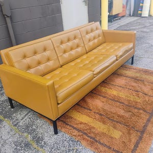 May include: A mustard yellow three-seat sofa featuring a tufted backrest and seat cushions. The sofa has a rectangular frame with straight armrests and is supported by four thin black metal legs, providing a structured and elevated appearance.
